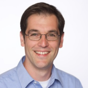 Professional headshot of Charlie Nicholls, a smiling man with short brown hair and glasses wearing a light blue shirt, against a plain light background.