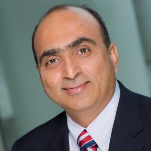 Professional headshot of Ravinder Dhawan, a smiling man in a dark suit, white shirt and striped tie, against a blurred background.