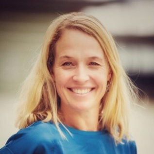 Professional headshot of Roos Steevels, a smiling woman with long blonde hair wearing a blue top, against a softly blurred outdoor background.