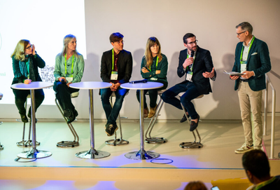 Several panellists seated on stools around small tables on a stage while a moderator stands holding a microphone at the GetReal Institute Conference 2024.