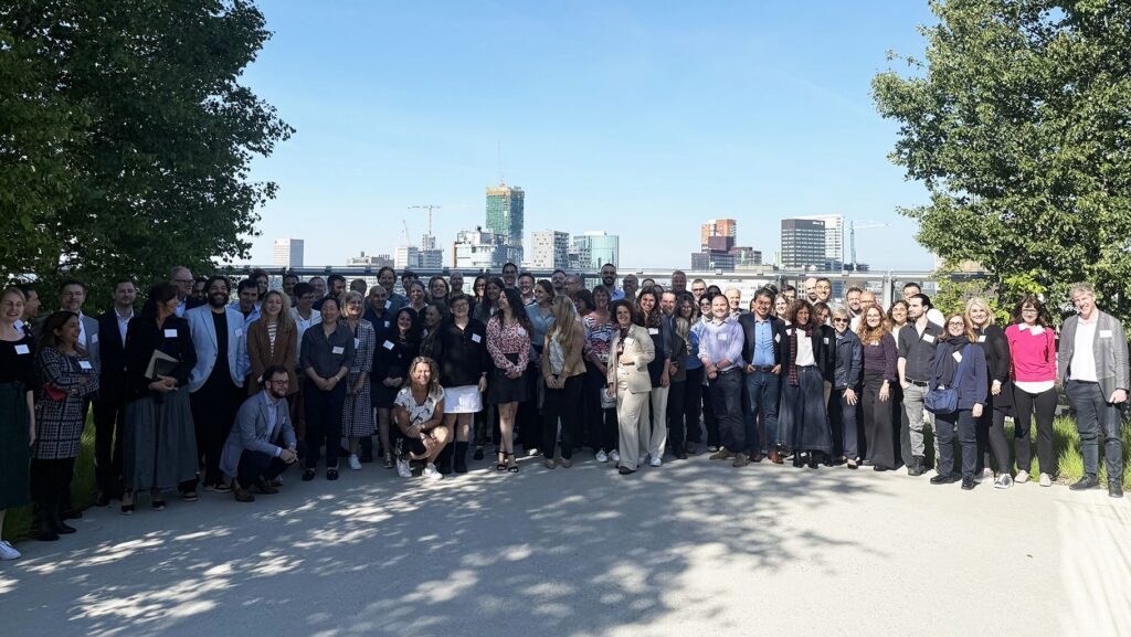 A large group of professionals wearing name badges posing together outdoors on a rooftop terrace with a city skyline behind them, at the official launch of GREG IHI.
