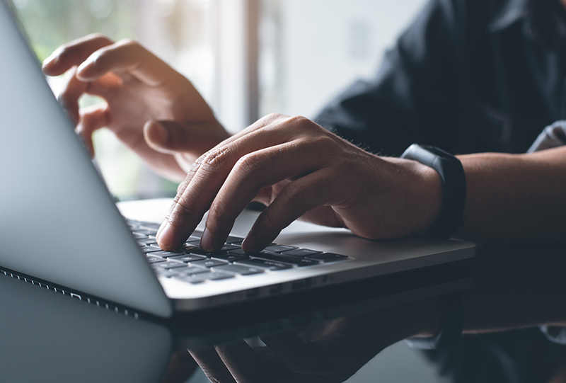 Close-up of a person's hands typing on a laptop keyboard near a window.