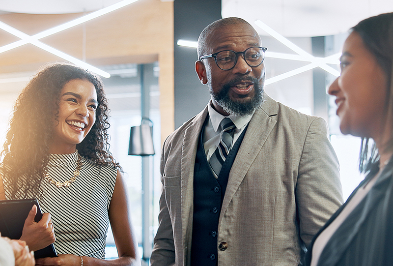 Three professionals smiling and talking together in a modern office: a man in a suit with glasses and a grey beard between two women.