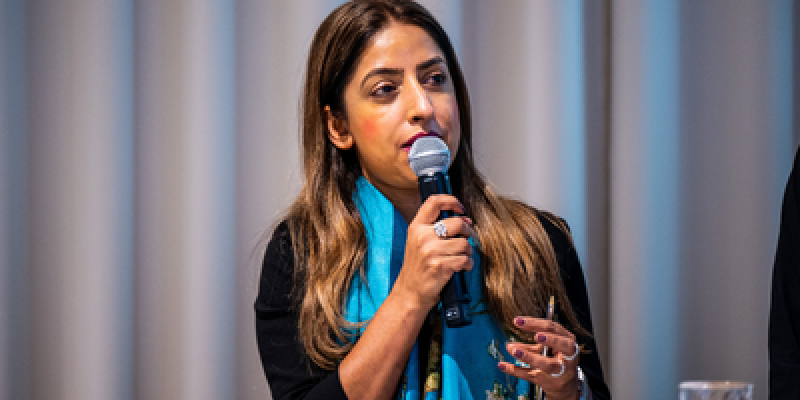 A woman with long dark hair wearing a turquoise scarf speaks into a handheld microphone and gestures at the GetReal conference.